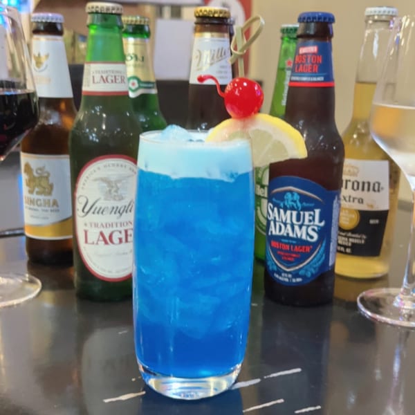 A vibrant blue cocktail with a cherry and lemon garnish, surrounded by various beer bottles.