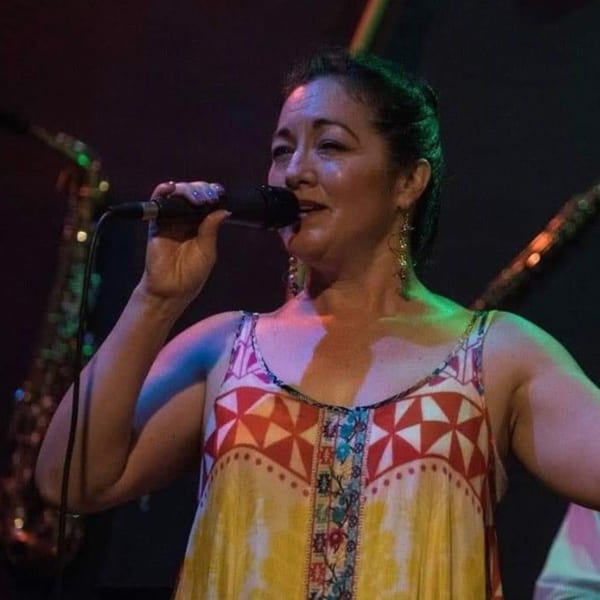 A woman in a colorful dress sings into a microphone on a dimly lit stage.