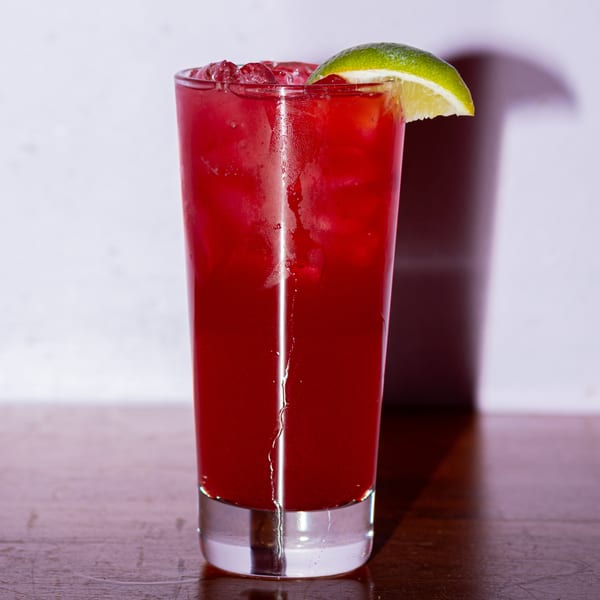 A tall glass of vibrant red Sala Thai iced drink with a lime wedge on a wooden table.