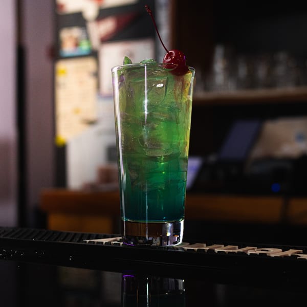 A vibrant green and blue cocktail with ice and a cherry on top, sitting on a bar.