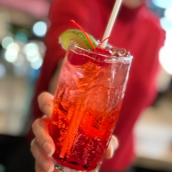 A hand holds a tall glass of red Sala Thai iced drink with lime, cherries, and a straw.