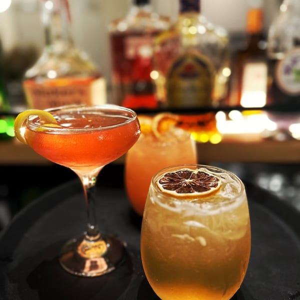 Three vibrant cocktails, one garnished with a lemon peel, on a dark tray with blurred liquor bottles in the background.