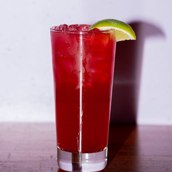 A tall glass of red iced drink, garnished with a lime wedge, on a wooden surface.