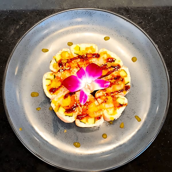 Flower-shaped sushi rolls with mango, cucumber, sauce, and a pink orchid on a gray plate.