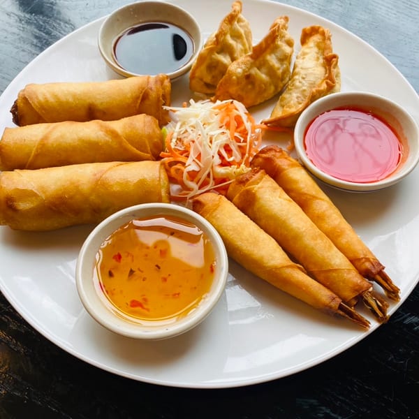 A white plate with Sala Thai spring rolls, dumplings, shrimp rolls, and dipping sauces.