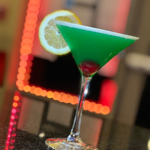 Green martini with a lemon slice and cherry, set against a blurred background of red lights.