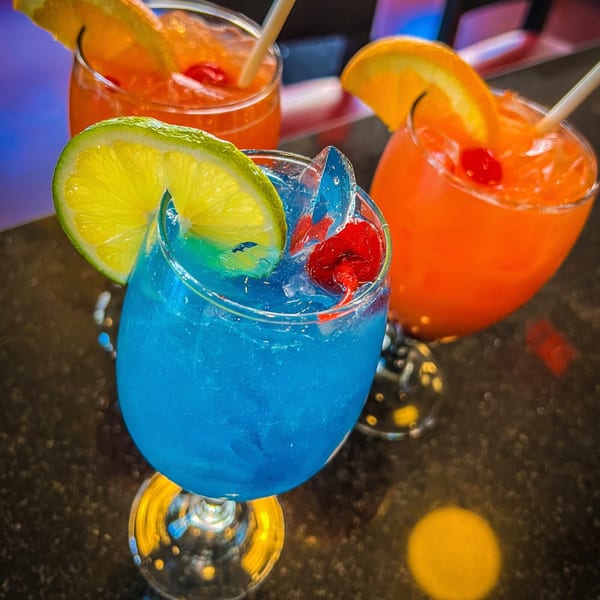 Three vibrant cocktails, one blue with lime, two orange with orange slices and cherries, on a dark surface.