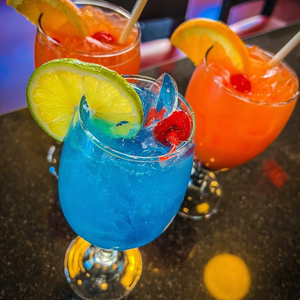 Three vibrant cocktails, one blue with lime, two orange with orange slices and cherries, on a dark surface.