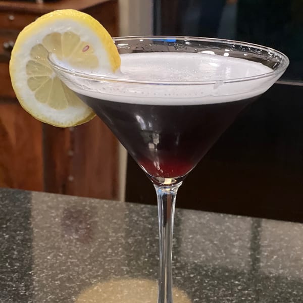 A dark cocktail with a white foamy top and a lemon slice garnish in a martini glass.