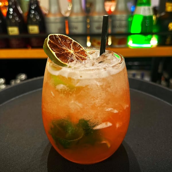 A vibrant orange cocktail with ice, a black straw, and a dried lime slice garnish.