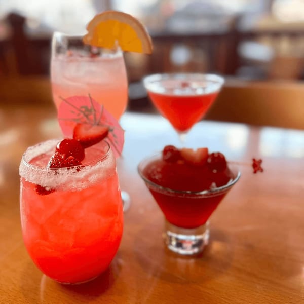 Three vibrant pink and red cocktails, two garnished with fruit and one with a small umbrella, on a wooden table.