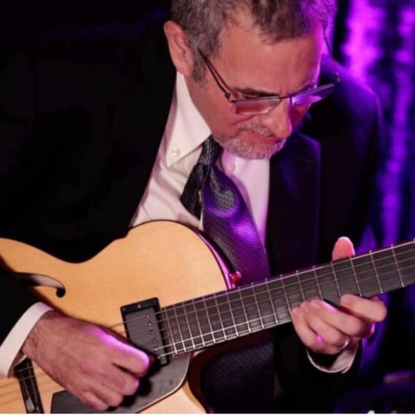 Man in a suit and tie playing an acoustic guitar under purple lighting.