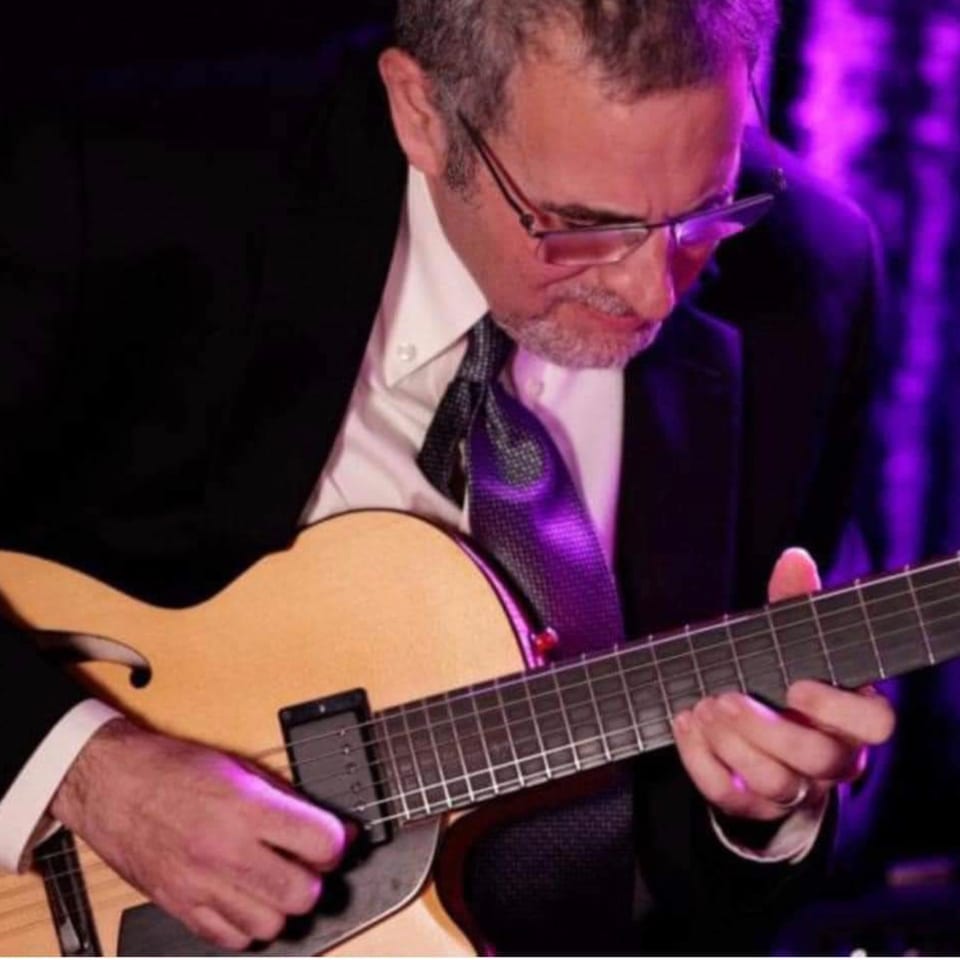 Man in a suit and tie playing an acoustic guitar under purple lighting.