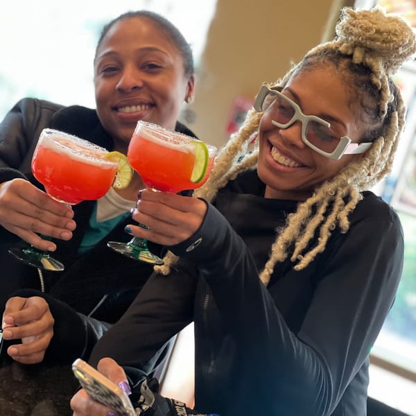 Two smiling women holding up bright pink margaritas with lime wedges and salt rims.