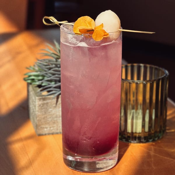 A tall glass of pink Sala Thai cocktail, garnished with lychee and dried orange slices.