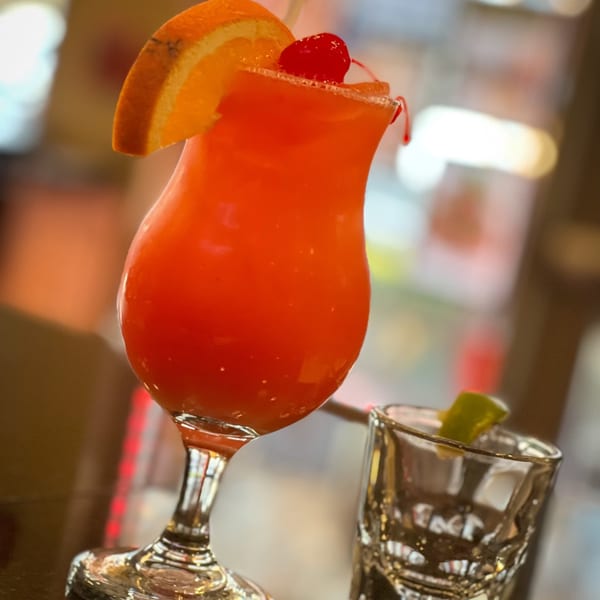 A vibrant orange cocktail garnished with an orange slice and cherry, next to a shot glass.