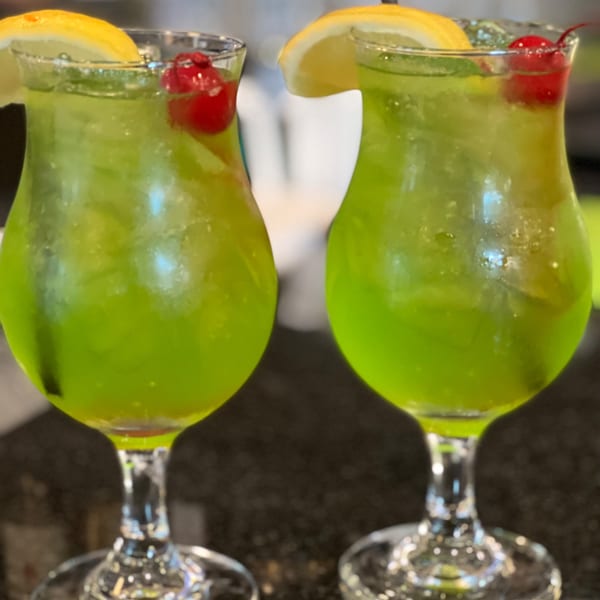 Two vibrant green Sala Thai cocktails, garnished with lemon slices and cherries, on a dark surface.