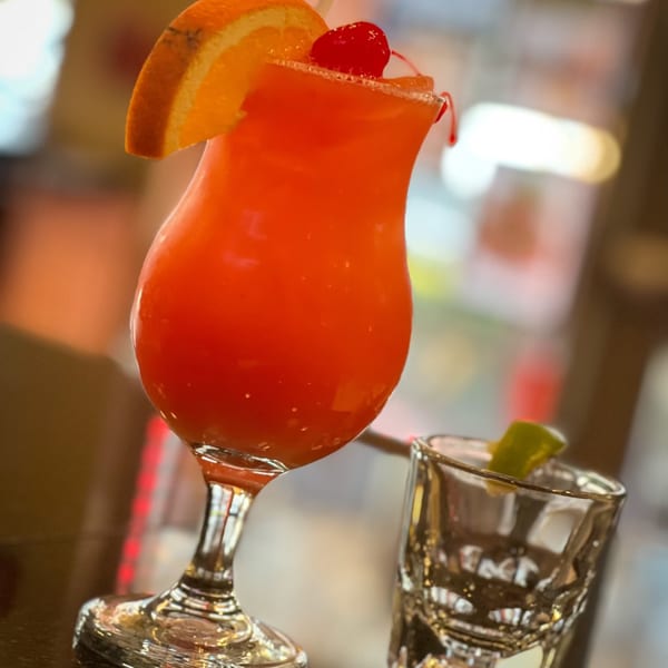 A vibrant orange cocktail with an orange slice and cherry garnish next to a shot glass.