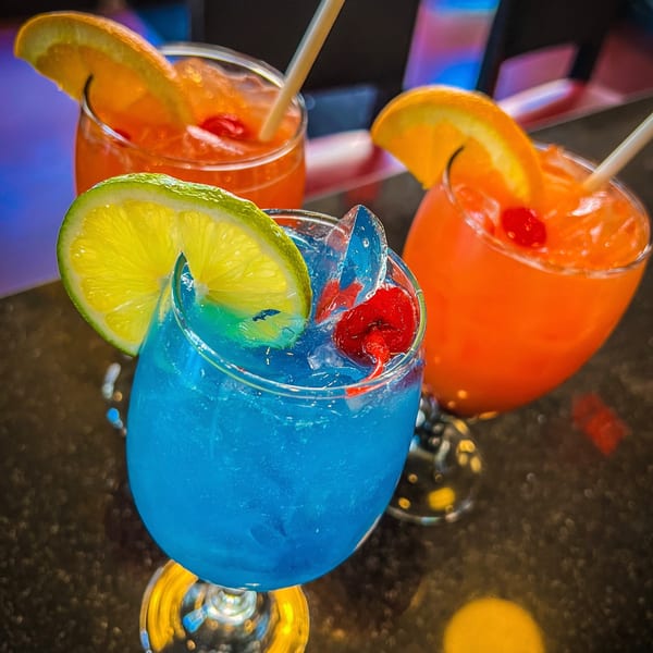 Three vibrant cocktails, one blue with lime, two orange with orange slices and cherries, on a dark surface.