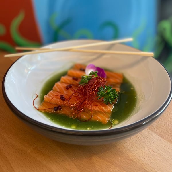 Salmon sashimi in green sauce with chili threads, microgreens, and a purple flower in a white bowl.
