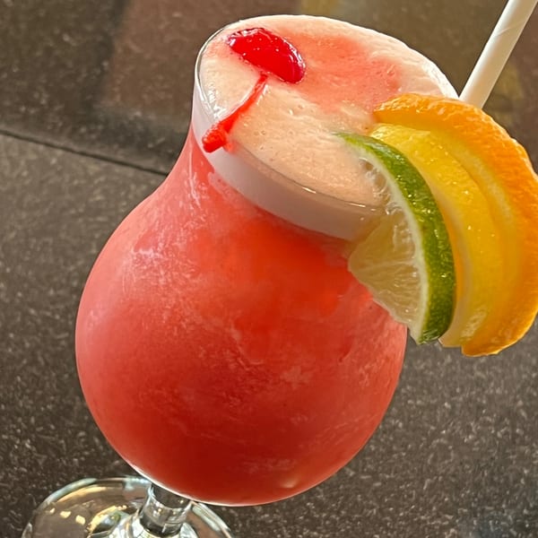 A vibrant red frozen cocktail, garnished with a cherry, lime, and orange slices, with a straw.