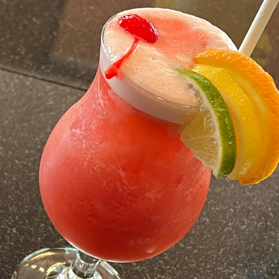 A vibrant red frozen cocktail, garnished with a cherry, lime, and orange slices, with a straw.