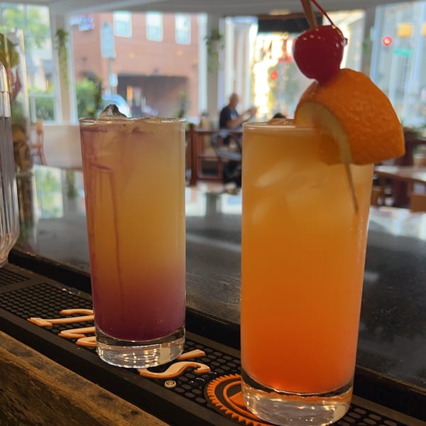 Two colorful Sala Thai cocktails, one purple and one orange, on a bar with a blurred background.