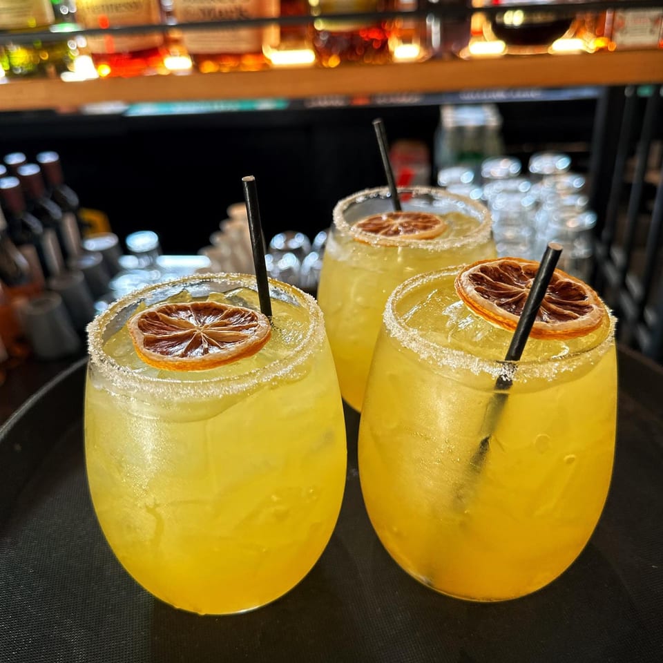 Three yellow cocktails with sugared rims, dried orange slices, and black straws on a dark tray.